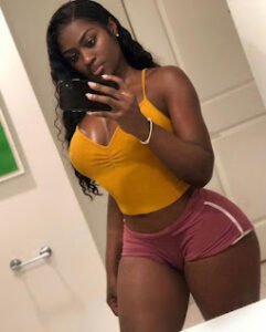 Curvy Black woman in mustard crop top and pink shorts taking mirror selfie, posing with seductive smile – Camille Blaze, UK adult phone chat model