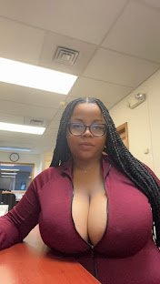 Busty woman with glasses in zipped maroon top, long braids, sitting at office desk with seductive expression – Nova Brooks, UK adult chat girl
