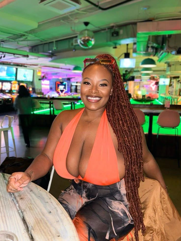 Busty British Jamaican woman in an orange top sitting at a bar in London with a radiant smile and long braids – Lola London, adult chat queen.