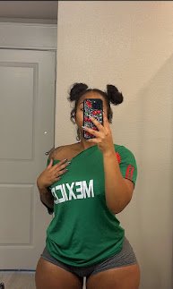 Thick Latina woman in green Mexico shirt and grey shorts taking mirror selfie with playful pose – Lola Fuego, UK adult phone chat girl