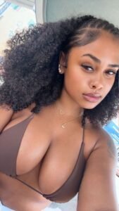 Busty Afro-Caribbean woman with curly hair, full lips, and seductive gaze wearing a brown bikini top, perfect for an adult phone chat profile from London.