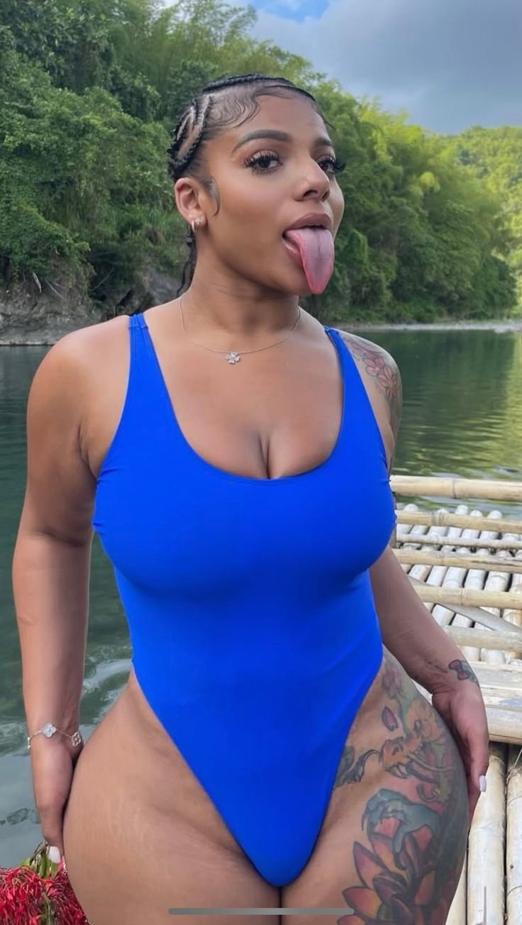 Curvy Caribbean girl in a royal blue swimsuit with tattoos, braids, and tongue out, standing on a bamboo raft near a river in Jamaica – Sasha Blu.