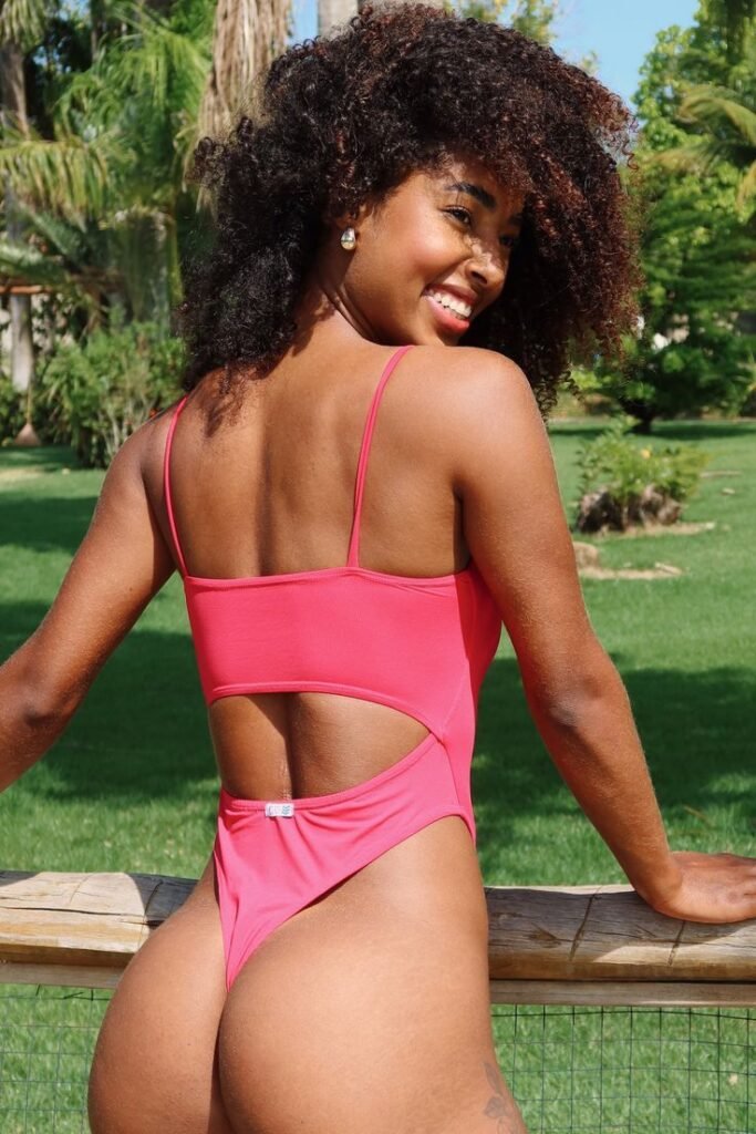 Curvy Caribbean woman in pink thong swimsuit, smiling flirtatiously outdoors – adult phone chat model in Brighton, UK.