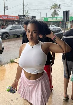 Curvy woman in tight wet white t-shirt and pink skirt standing outside in rain, holding her hair with a flirty smile – UK adult phone chat tease Zari Belle