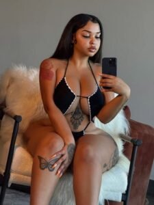 Curvy tattooed Caribbean woman in revealing black bikini, sitting on a furry chair taking a sultry mirror selfie – perfect for seductive phone chat.