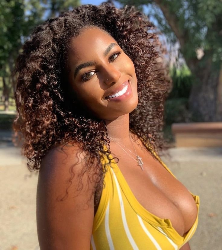 Curvy Caribbean woman with golden smile and deep neckline, wearing a yellow dress in sunlight, perfect for seductive phone chat profile.