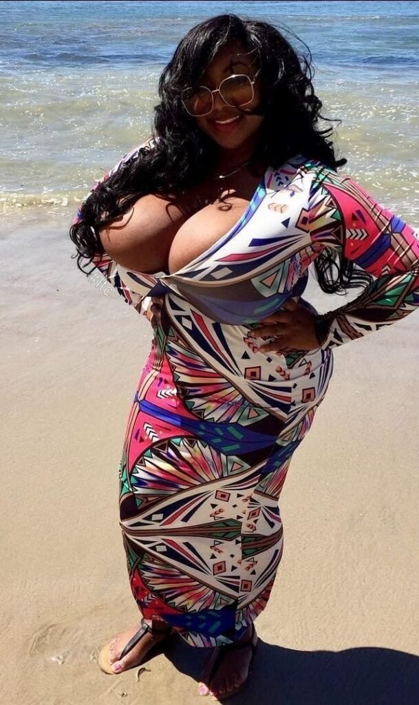 Busty black woman wearing a colorful body-hugging beach dress standing on a sunny UK beach – Miss Coco, adult voice chat operator in Brighton.