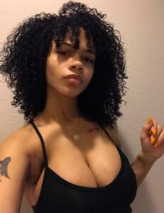 Curvy woman with jet black curly hair, full cleavage, and confident expression posing in a black top for an adult phone chat profile.