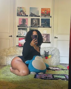 Thick woman kneeling in front of mirror on green rug, curly hair, black bodysuit, plush toys and album wall decor – UK adult chat girl Nia Velvet