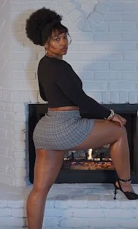 Tall Black woman posing in plaid mini skirt and heels by fireplace, afro puff and seductive stare – Zahara Stone, UK adult phone fantasy tease