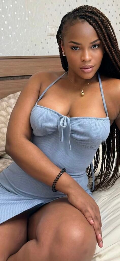 Curvy Caribbean woman in blue dress sitting on a bed – adult phone chat in Manchester.