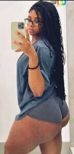 Mirror selfie of curvy Caribbean woman with long braids and glasses wearing a grey top and shorts – Kayla Storm, adult phone chat girl in UK