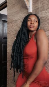 Beautiful Afro-Caribbean woman in a tight red dress with long black locs, posing confidently in a doorway – perfect for a sexy UK voice chat profile.