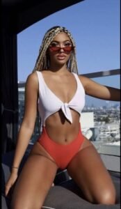 Light-skinned woman in a red and white swimsuit, braided hair and sunglasses, posing confidently on a rooftop in Manchester – adult phone chat model.