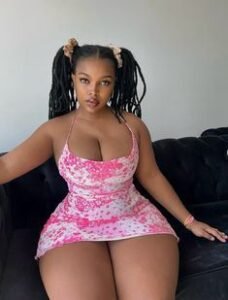 Curvy Caribbean girl with long twin ponytails wearing a tight pink dress, sitting on a sofa with a seductive look – Princess Kandy from Trinidad.