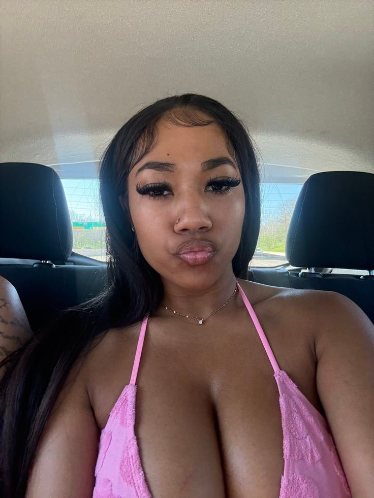 Busty Caribbean-British woman in pink top, pouting seductively in a car – adult phone chat in London profile image.