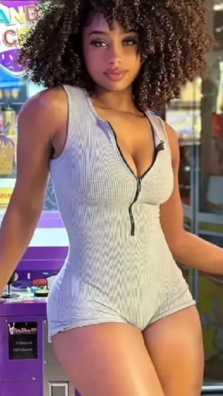 Curvy Caribbean girl with natural curly hair in a zip-up grey romper, posing in front of a game machine — Imani Rose, adult phone chat in Leeds
