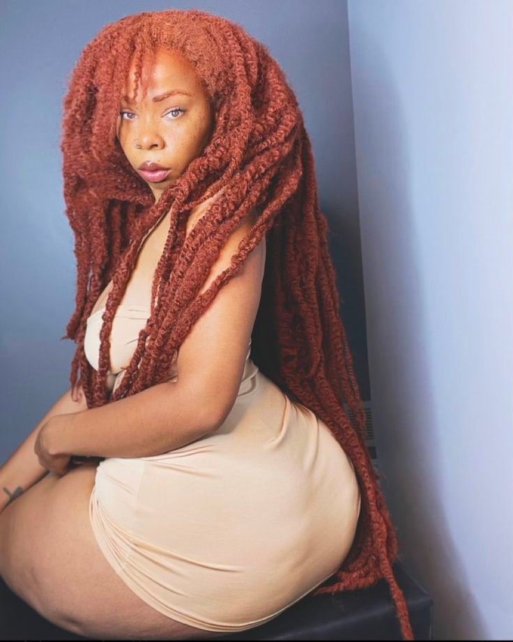 Caribbean beauty Aaliyah Ember with long red locs, wearing a beige mini dress and giving a sultry side look while sitting.