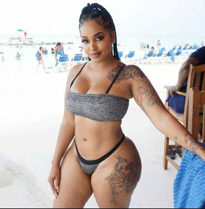 Grenadian beauty Zaria Vibe in a grey bikini, standing on the beach with tattoos on her arms and hip, smiling with a sultry gaze
