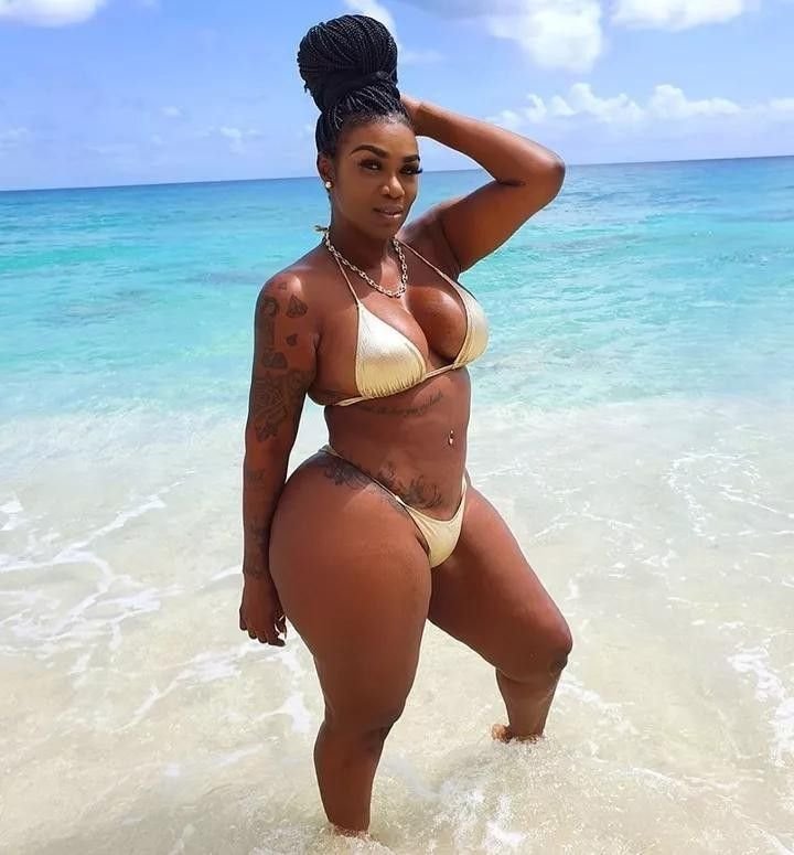 Caribbean beauty Tiana Gold in a gold bikini, standing in the shallow turquoise water with one hand on her head, looking confidently at the camera in London-inspired beach shoot style