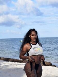 Caribbean beauty Keira Flame with long wavy black hair, wearing a cropped white top and black bikini bottoms, standing on the beach with waves in the background.