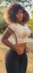 Trinidadian beauty Amara Storm with curly hair in a tight white crop top and jeans, posing outdoors with a sultry gaze.