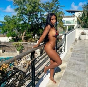 Barbadian beauty Sienna Blaze in a tiny black bikini, leaning against a balcony with ocean and trees in the background.