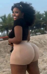 Caribbean beauty Liora Shade with natural afro hair, wearing a black top and beige shorts, smiling over her shoulder on a sandy beach.