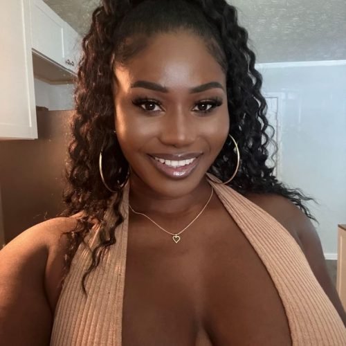 Phone Chat Seductress in Manchester Naomi Blackwood – busty Caribbean woman with curly hair, smiling seductively in a low-cut top.