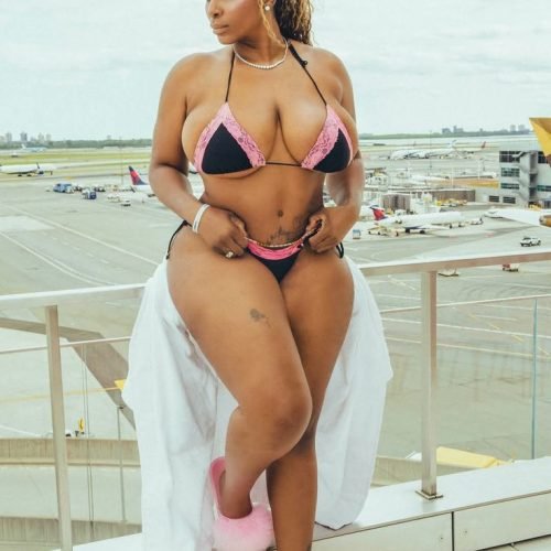 Jamaican beauty Layla Luxe in a black and pink bikini with a white cover-up, posing on a balcony overlooking an airport runway