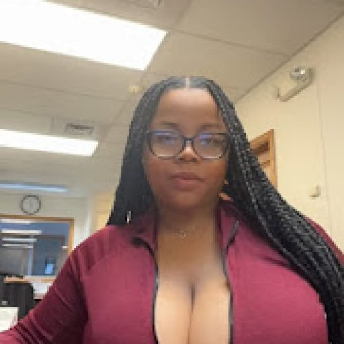 Busty woman with glasses in zipped maroon top, long braids, sitting at office desk with seductive expression – Nova Brooks, UK adult chat girl