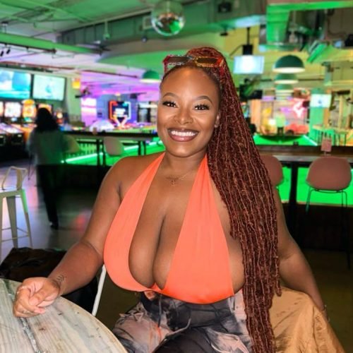 Busty British Jamaican woman in an orange top sitting at a bar in London with a radiant smile and long braids – Lola London, adult chat queen.