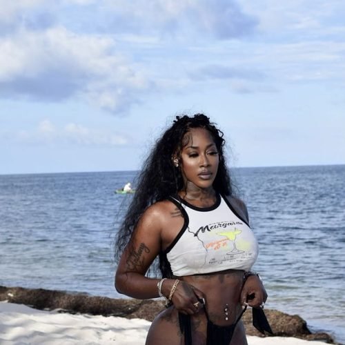 Caribbean beauty Keira Flame with long wavy black hair, wearing a cropped white top and black bikini bottoms, standing on the beach with waves in the background.