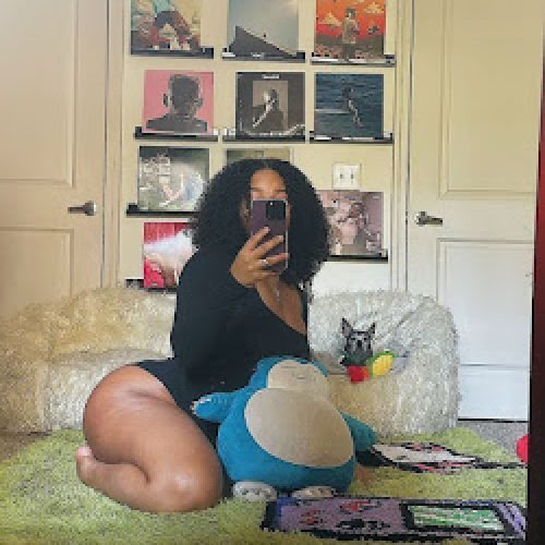 Thick woman kneeling in front of mirror on green rug, curly hair, black bodysuit, plush toys and album wall decor – UK adult chat girl Nia Velvet