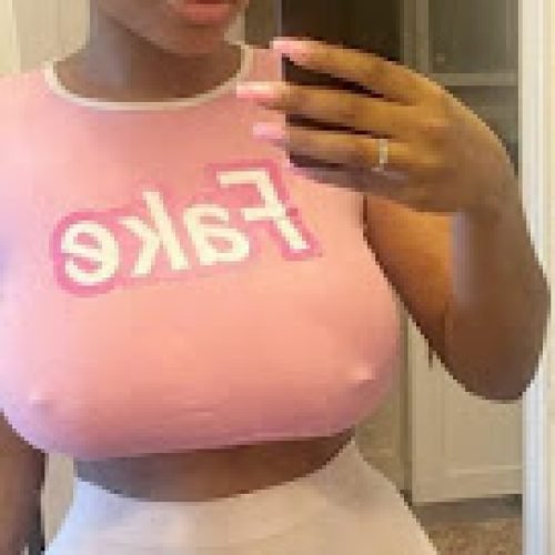 Curvy woman in pink crop top that says “fake” and white sheer shorts taking a mirror selfie – Trixie Starr, UK adult phone chat tease