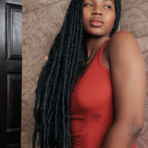 Beautiful Afro-Caribbean woman in a tight red dress with long black locs, posing confidently in a doorway – perfect for a sexy UK voice chat profile.