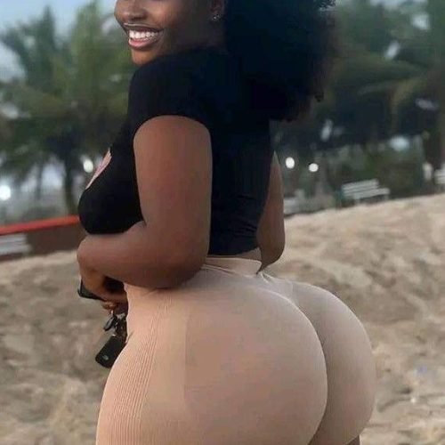 Caribbean beauty Liora Shade with natural afro hair, wearing a black top and beige shorts, smiling over her shoulder on a sandy beach.